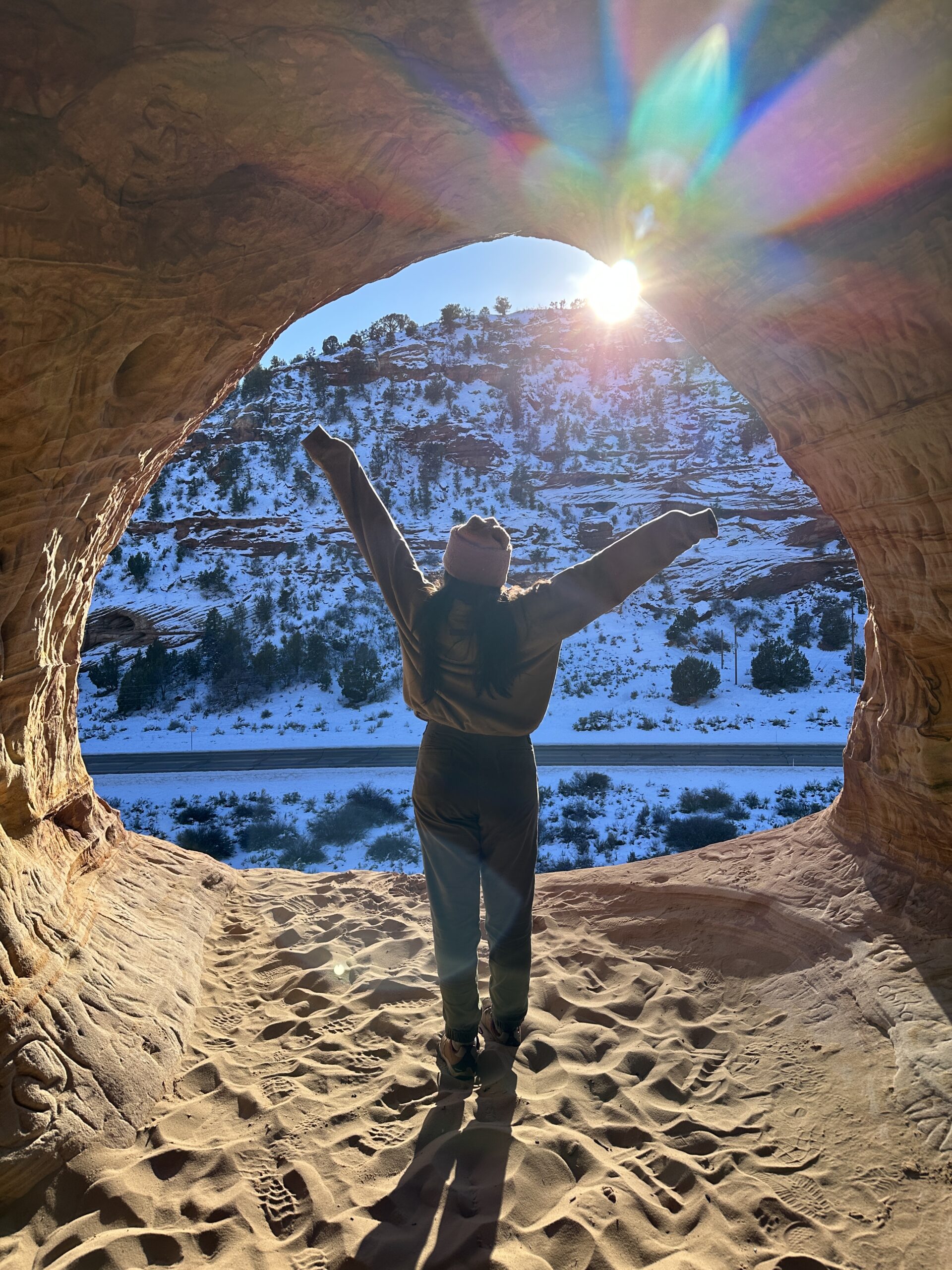 A person stands at a cave entrance, facing the mountains opposite.