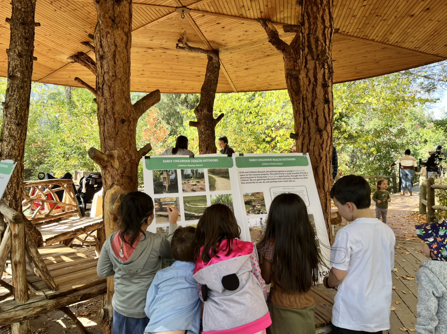 Under a pavilion at a park, five small children look at a presentation poster board. There are other children and adults around.