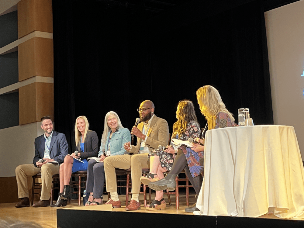 Six people sit on stage. One person is holding a microphone and speaking.