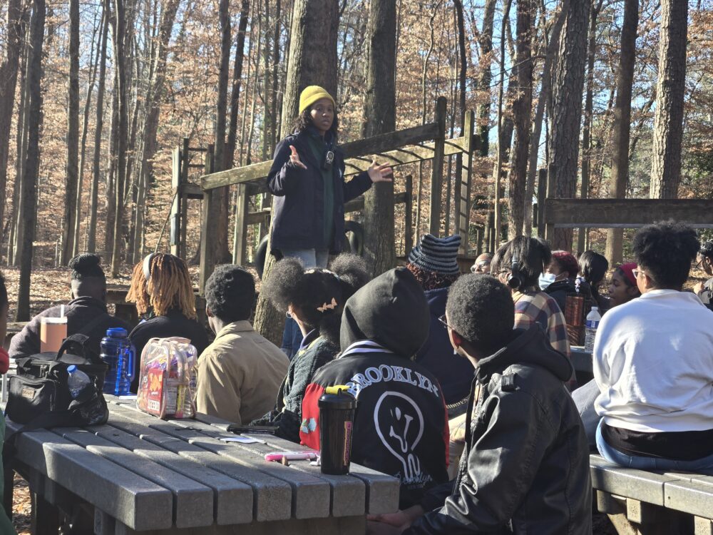 One adult stands before a group of students in the woods, speaking with them.